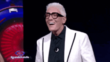a man in a white suit and glasses is laughing in front of a sign that says grande fratello