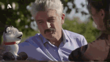a man with a mustache is talking to a woman while holding a dog