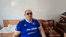 a man wearing sunglasses and a blue shirt that says mw dias ostrava sits on a bed