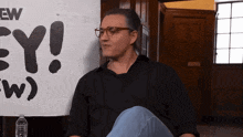 a man wearing glasses sits in front of a sign that says ew