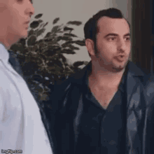 two men are standing next to each other in a room . one of the men is wearing a leather jacket .