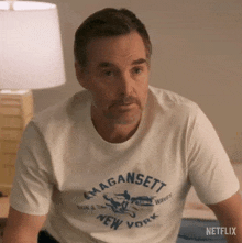 a man wearing a white t-shirt is sitting on a bed and looking at the camera .