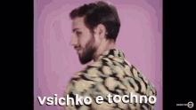 a man with a beard is standing in front of a pink background . he is wearing a colorful shirt .