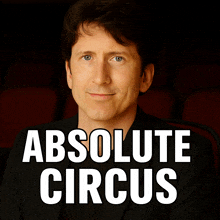 a man in a black suit sits in front of a poster for absolute circus