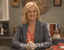 a woman in a suit is sitting at a desk and smiling with the words `` naturally '' written on her face .