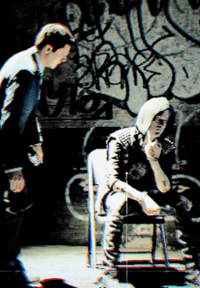 a man standing next to a woman sitting in front of a wall with graffiti on it that says ' i love you '