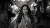 a black and white photo of a woman in a trench coat walking down a street at night .