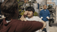 a man in a blue jacket is standing behind a man in a maroon hoodie who is fighting another man