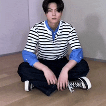 a young man wearing a striped shirt and jeans is sitting on the floor .