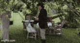 a man is standing in a garden reading a book while a woman looks on .