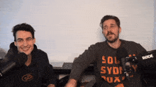 a man wearing a south sydney sweatshirt stands next to another man wearing a rode microphone