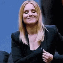 a woman in a black jacket and black top is smiling and holding her hands together .