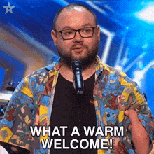 a man wearing glasses is standing in front of a microphone and saying what a warm welcome !
