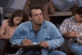 a man in a blue jacket is sitting at a desk in a classroom with other people .