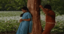 a man and a woman are standing next to a tree . the woman is wearing a blue sari .