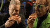 two men are eating slices of pizza and one is wearing green glasses