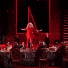 a drag queen in a red dress is dancing on a stage with her arms outstretched .