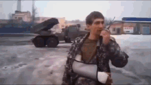 a man with a cast on his arm is talking into a microphone in front of a military vehicle .