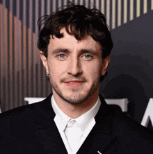 a man with curly hair and a beard wears a black suit