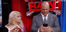 a man in a suit and tie is looking at his cell phone next to a woman in a wrestling championship belt .