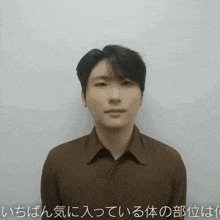 a man wearing a brown shirt is standing in front of a white wall with foreign writing on it