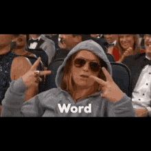 a woman wearing a hoodie and sunglasses is sitting in a crowd of people and making a peace sign .