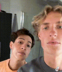 two young men are standing next to each other and looking at the camera . one of the men is wearing an orange shirt .
