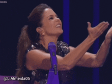 a woman applauds in front of a purple microphone with the username lialmeida05