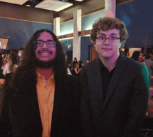 a man with long hair and glasses stands next to another man in a suit