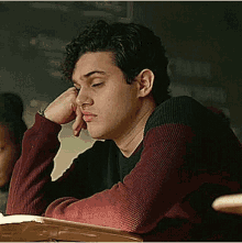a young man is sitting at a desk in a classroom with his head resting on his hand .