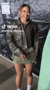 a woman wearing a green skirt and a leather jacket is standing in front of a black tarp with tik tok written on it
