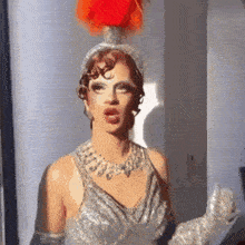 a drag queen wearing a silver dress and gloves has a red feather on her head .