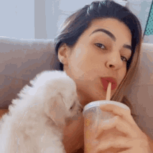 a woman is drinking from a cup with a straw while holding a small white dog .