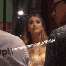 a woman in a white dress is talking to a man in a black shirt that says emmamuscat official