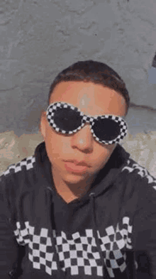 a young man wearing a black and white checkered shirt and sunglasses is looking at the camera .