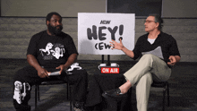 two men are sitting in front of a sign that says hey
