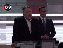 two men are standing in front of a clock that says 09
