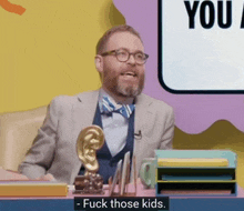 a man in a suit and bow tie is sitting at a desk with a sign that says " you " behind him