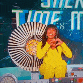 a woman in a yellow outfit is making a heart shape with her hands while sitting in front of a clock that says time