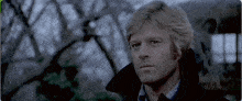 a man with blonde hair is wearing a black trench coat .