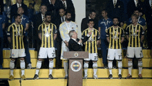 a man stands at a podium in front of a group of soccer players one of whom has the number 25
