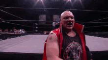 a bald wrestler in a red vest is standing in the middle of a wrestling ring .