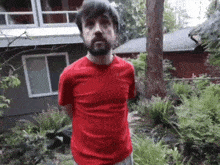 a man with a beard wearing a red shirt is standing in front of a house