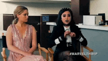 two women are sitting in chairs in an office . one is wearing a pink dress and the other is wearing a black jacket .