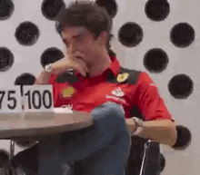 a man in a red shirt is sitting at a table with a sign that says 75,000 on it .