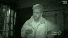 a man in a white jacket is standing in a dark room with travel channel written on the bottom of the screen .