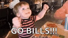 a little boy is sitting at a table in a restaurant with his fist in the air and saying `` go bills '' .