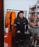 a man in a black jumpsuit is dancing in a room with orange jackets on a rack