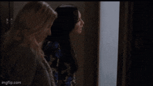 two women are standing next to each other in a room looking out of a window .