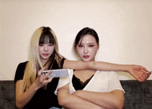 two women are sitting on a couch with their arms crossed and one is holding a cell phone